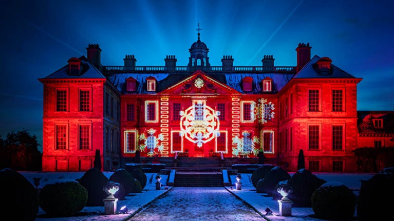 Belton House is lit up in red with yellow and white snowflakes across it. The gardens are in the foreground and there is a scattering of snow on the ground.